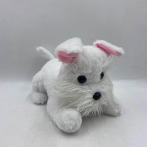 Puede incluir: Un perro de peluche blanco con orejas rosas y ojos negros. El perro está acostado con la cabeza girada hacia un lado.