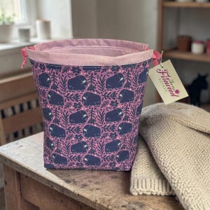 May include: A fabric storage basket with a pink interior and a drawstring closure. The exterior features a pattern of dark blue bison and foliage on a mauve background. A tag with the text "Fireweed Studio" is attached.