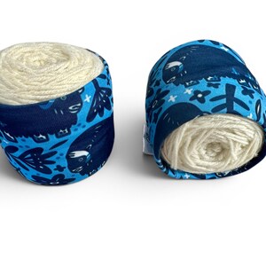May include: Two yarn bowls, each holding a ball of cream-colored yarn. The bowls are wrapped in blue fabric with a pattern of dark blue animals, flowers, and leaves. The bowls are designed to keep yarn clean and prevent tangling.