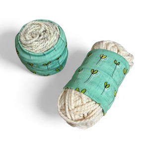 May include: Two skeins of off-white yarn are shown, one in a teal fabric container and the other wrapped with a teal band. The teal fabric is patterned with green sprouts and white stems. The yarn appears to be a bulky weight.
