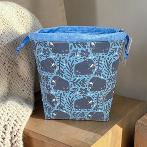 May include: A square fabric storage basket with a blue denim interior and drawstring closure. The exterior features a light blue background with a pattern of dark blue musk oxen and floral elements. A tag is attached to the side.