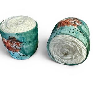 May include: Two spools of white yarn are displayed. Each spool is wrapped in a teal fabric with a watercolor-style illustration of a brown and orange animal. The yarn is tightly wound, and the fabric has a speckled pattern.