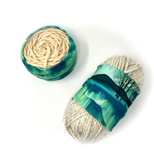 May include: Two balls of yarn, one in a green and blue fabric cover with a white yarn ball inside, and the other in a green and blue fabric cover with a white yarn ball on the outside.