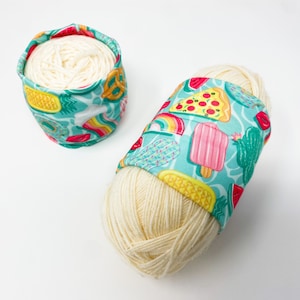 May include: Two balls of cream-colored yarn wrapped in turquoise fabric with a colorful print of summer food like watermelon, pizza, ice cream, and pineapple.