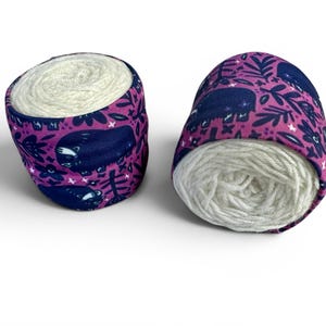 May include: Two yarn bowls, each holding a ball of white yarn. The bowls are covered in a purple fabric with a pattern of dark blue bears, leaves, and flowers. The bowls are on a white surface.