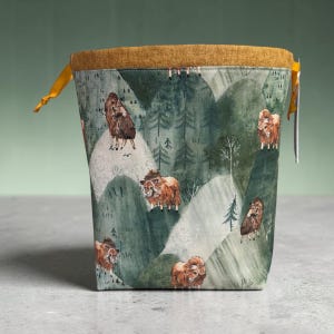 May include: A fabric project bag with a mustard yellow rim and drawstring. The bag features a repeating pattern of brown musk oxen on a green and white mountain landscape. The bag is designed to hold knitting or crochet projects.