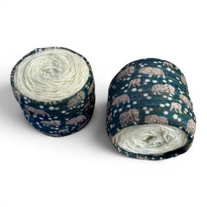May include: Two spools of yarn. Each spool is wrapped in a dark teal fabric with a pattern of sheep and white star-shaped accents. The yarn itself is a creamy white color.
