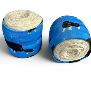 May include: Two yarn bowls, each holding a ball of cream-colored yarn. The bowls are cylindrical, with a blue fabric exterior featuring a pattern of black and white dogs and snowflakes. The yarn is visible at the top.