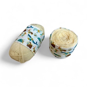 May include: Two skeins of yarn. One is a cylindrical shape with a patterned fabric band featuring various animal illustrations. The other is a round container filled with yarn. The yarn is a light beige color, and the fabric band has a white background with blue, brown, and teal accents.