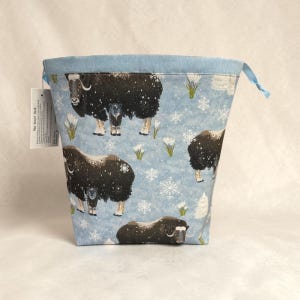 May include: A light blue drawstring bag with a repeating pattern of black musk oxen, snowflakes, and white flowers. The bag has a light blue rim and a blue drawstring. The tag reads "The Scarf Sock".