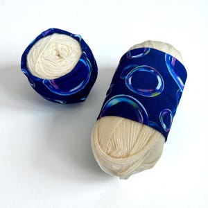May include: Two balls of cream-colored yarn are shown, one inside a blue fabric bowl and the other wrapped with a blue fabric band. The fabric has a pattern of large, colorful bubbles.