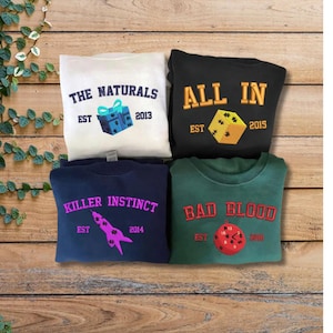 Può includere: Quattro felpe con disegni ricamati. La felpa bianca dice "THE NATURALS" con un'immagine di confezione regalo. La felpa nera dice "ALL IN" con un'immagine di dado. La felpa blu navy dice "KILLER INSTINCT" con un'immagine di razzo. La felpa verde dice "BAD BLOOD" con un'immagine di orologio.