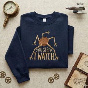 Puede incluir: Sudadera azul marino con un diseño bordado marrón de una criatura mecánica y el texto "YOU SLEEP. I WATCH." La sudadera está doblada, con accesorios de estilo steampunk.