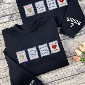 Puede incluir: Dos sudaderas negras con diseños bordados. Cada una presenta una serie de parches tipo sello postal con frases e imágenes. Una manga tiene "GIBSIE 7" y la otra "KAVANAGH 13". Las sudaderas tienen cuello redondo.