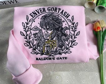 May include: Pink sweatshirt with a black and gold embroidered design featuring a character named "Enver Gortash" from the game "Baldur's Gate".