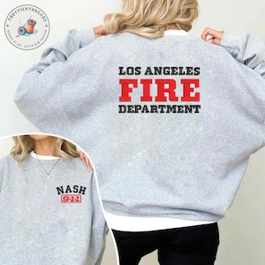 Può includere: Una felpa grigio chiaro con la scritta "LOS ANGELES FIRE DEPARTMENT" in rosso e nero sul retro. La parte anteriore della felpa presenta la scritta "NASH 911" in nero e rosso. La felpa ha un colletto bianco.