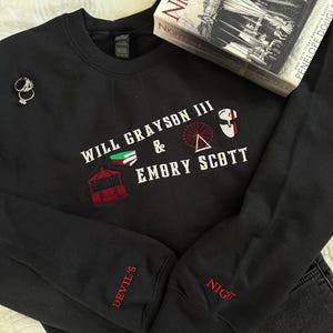 May include: A black sweatshirt with embroidered text and graphics, including "WILL GRAYSON III & EMBRY SCOTT." The sleeves have "DEVIL'S" and "NIGHT" in red. A book titled "NIGHTFALL" and a pair of rings are also visible.