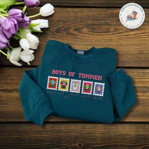 May include: Teal sweatshirt with the text "BOYS OF TOMMEN" in pink. Below the text are five postage stamp designs with jersey numbers. The sweatshirt is folded on a wooden surface, with flowers in the upper left corner.