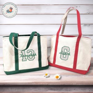Puede incluir: Dos bolsas de lona con texto y números bordados. Una bolsa tiene ribetes y asas verdes con "13 Savannah" en verde. La otra tiene ribetes y asas rojas con "6 Lynch" en verde. Ambas bolsas están sobre una superficie de madera.