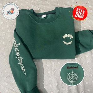 Puede incluir: Una sudadera verde oscuro con bordado blanco de una espada con espinas en una manga y un conjunto de colmillos en la parte delantera. La sudadera tiene el texto "Thuytienthreads" en el hombro izquierdo y "Behind" en el lado derecho.