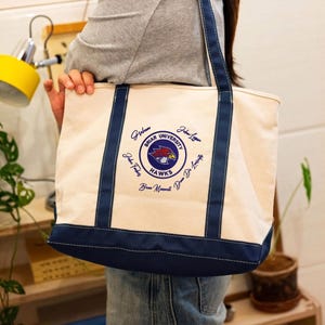 Puede incluir: Bolso tote de lona color crema con ribete y asas azul marino. La bolsa presenta un emblema circular con un halcón rojo y dorado, las palabras "Briar University Hawks" y varias firmas en azul marino.