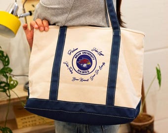 May include: A cream-colored canvas tote bag with dark blue trim and handles. The center features a circular logo for Briar University Hawks with a red and blue bird mascot, surrounded by several handwritten names in blue ink script.