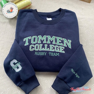 Puede incluir: Sudadera azul marino con el texto "TOMMEN COLLEGE RUGBY TEAM" en verde y blanco. La manga izquierda tiene el número "6" y la palabra "Lunch" bordada en gris. La manga derecha tiene las palabras "Nice legs" bordadas en verde.
