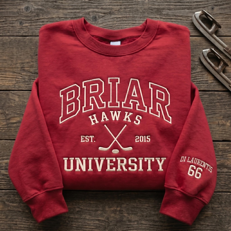K&ouml;nnte beinhalten: Ein rotes Sweatshirt mit wei&szlig;em Schriftzug "BRIAR HAWKS UNIVERSITY". Das Design umfasst gekreuzte Hockeyschl&auml;ger und das Jahr "2015". Auf dem &Auml;rmel stehen der Name "Di Laurentis" und die Nummer "66".