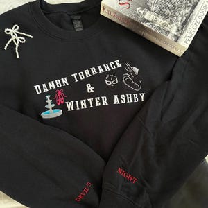 Puede incluir: Sudadera negra con texto bordado blanco "DAMON TORRANCE & WINTER ASHBY". Presenta una fuente y zapatillas de ballet. La manga tiene "DEVIL'S" y "NIGHT" en rojo. Un libro titulado "Kill Switch" está encima.