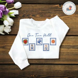 May include: A white long-sleeve sweatshirt featuring the blue text One Tree Hill above five postage stamp-style graphics. The icons include a flaming heart, a basketball player in a jersey, a raven, another player in a jersey, and a basketball.