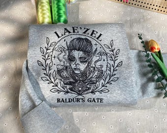 May include: A gray sweatshirt with a black embroidered design featuring a character named Lae'zel from the game Baldur's Gate. The design includes a circular frame with a floral border and the text "Lae'zel" and "Baldur's Gate".