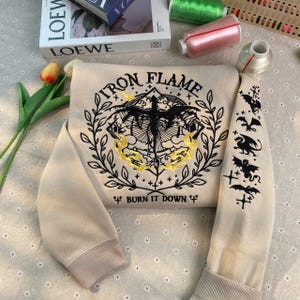 Puede incluir: Una sudadera beige con detalles bordados en negro. La parte delantera presenta un dragón con llamas y el texto "Iron Flame" y "Burn It Down". Las mangas tienen dragones y cruces bordados en negro.
