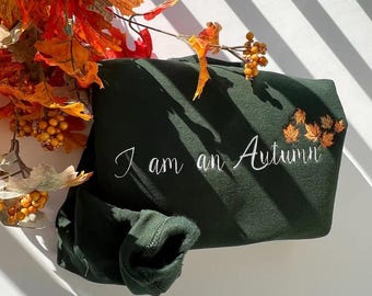 May include: Dark green sweatshirt with the embroidered phrase "I am an Autumn" in white script. The sweatshirt is adorned with embroidered orange leaves. Autumnal foliage, including orange and yellow leaves, is arranged around the sweatshirt.