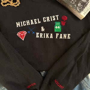 May include: Black long-sleeve sweatshirt with embroidered text and graphics. The text reads "MICHAEL CRIST & ERIKA FANE" with a diamond, a jersey, and a mask graphic. The sleeves have "DEVIL'S" and "NIGHT" embroidered in red. A gold chain and a book are also in the image.