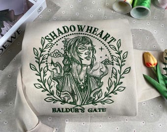 May include: A beige sweatshirt with a green embroidered design featuring a woman with a crown and the text "Shadowheart Baldur's Gate".