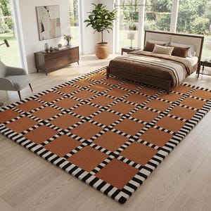 Op de afbeelding: Een rechthoekig vloerkleed met een geometrisch patroon. Het kleed heeft een raster van vierkanten in een warme bruine kleur, omrand met zwart-witte strepen. Het kleed bevindt zich in een slaapkamer en voegt een decoratief element toe.