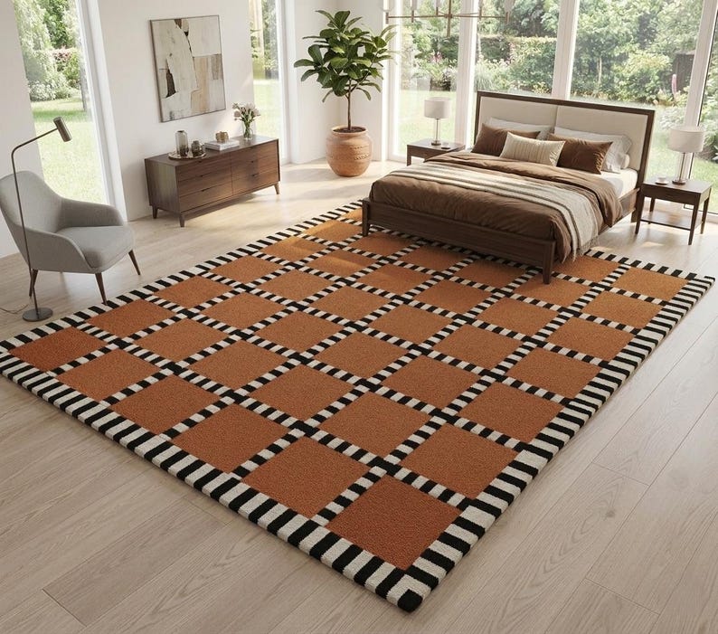 Op de afbeelding: Een rechthoekig vloerkleed met een geometrisch patroon. Het kleed heeft een raster van vierkanten in een warme bruine kleur, omrand met zwart-witte strepen. Het kleed bevindt zich in een slaapkamer en voegt een decoratief element toe.
