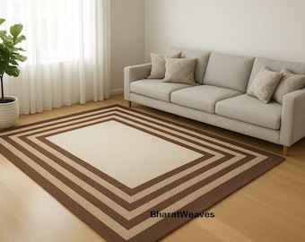Puede incluir: Una alfombra rectangular con un centro crema y un borde marrón. La alfombra está colocada sobre un suelo de madera frente a un sofá de color claro. Una planta en maceta está a la izquierda de la alfombra. Dimensiones aproximadas: 200 cm x 300 cm.