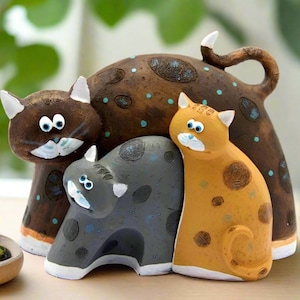 以下が含まれることがあります： 青い斑点のある茶色の猫、茶色の斑点のあるオレンジ色の猫、青い斑点のある灰色の猫の3つのセラミック製の猫の置物。猫たちは遊び心のあるポーズで配置されており、茶色の猫が後ろ、オレンジ色の猫と灰色の猫が前にいます。