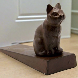 May include: A brown ceramic cat doorstop with a wedge-shaped base. The cat is sitting with its paws tucked under its body.