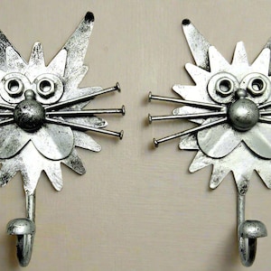 Può includere: Due ganci in metallo argentato a forma di gatto con un design fantasioso. Ogni gancio ha una testa rotonda con due grandi occhi rotondi e un naso rotondo più piccolo. I baffi del gatto sono fatti di chiodi di metallo.