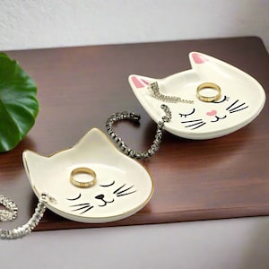 Può includere: Due portaoggetti in ceramica bianca a forma di gatto con bordo dorato. Ogni portaoggetti ha un viso di gatto nero con guance rosa e occhi chiusi. Un portaoggetti contiene un anello dorato e una catena d'argento, mentre l'altro contiene un anello d'argento e una catena nera.