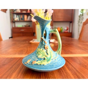 May include: A turquoise ceramic pitcher featuring a wide, flat base and a narrow neck with a single handle. The surface is decorated with embossed light green leaves and small brown berries, set against a textured, horizontal ridged pattern.