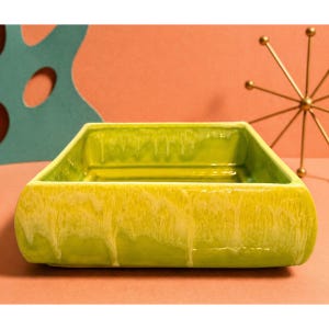 May include: A square, ceramic dish with a glossy, lime green glaze. The glaze has a dripped effect, with lighter yellow streaks running down the sides. The dish is empty and sits on a peach-colored surface, with a gold starburst decoration in the background.