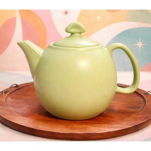 May include: A light green ceramic teapot with a rounded body, curved handle, and a lid with a small knob. The teapot sits on a round, wooden serving tray. The background features a colorful, retro-style pattern.