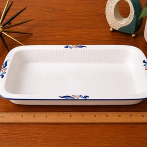 May include: A rectangular white ceramic baking dish with ribbed sides and a blue rim. The dish features blue leaf and orange berry motifs on the edges. A wooden ruler placed in front shows the dish is approximately 12.5 inches in length.