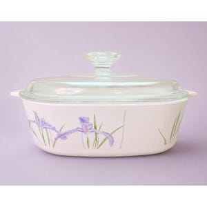 May include: An oval white casserole dish with a clear glass lid and a knob handle. The exterior features a painted pattern of purple iris flowers with green stems and leaves. The dish has integrated side handles and a smooth, glossy finish.
