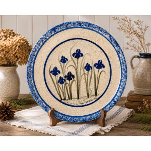 May include: A ceramic plate with a cream-colored center and a blue floral design. The plate has a blue rim with a speckled pattern and is supported by a wooden stand. The floral design features blue irises with green stems.