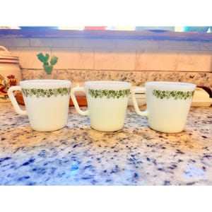 May include: Three white ceramic mugs with a green floral pattern around the rim. Each mug has a curved handle and a slightly flared shape. The mugs are displayed on a speckled countertop.