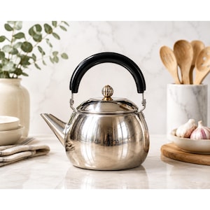 May include: A polished stainless steel tea kettle with a rounded body, a curved spout, and a black arched handle. The lid features a small, vertically ribbed metallic knob with gold-toned accents at the base, providing a reflective surface.
